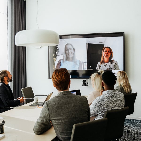 En grupp kollegor sitter i en konferensrum och har videomöte på två kollegor på stor skärm