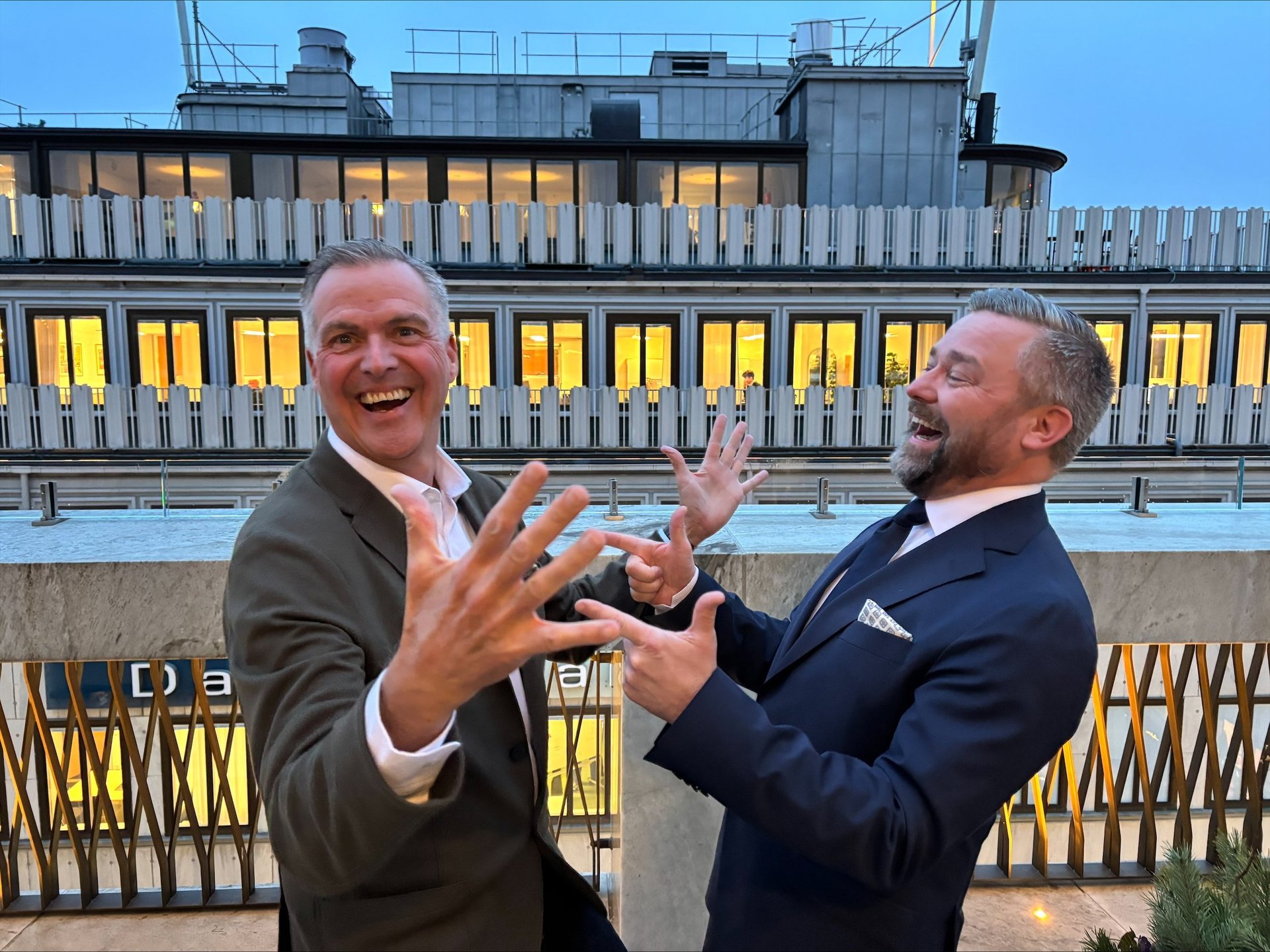 Tomas Wanselius and Joachim Westman står på en terrass framför en byggnad med upplysta fönster. De skrattar och gör stora gester med händerna, vilket skapar en lekfull och energisk stämning.