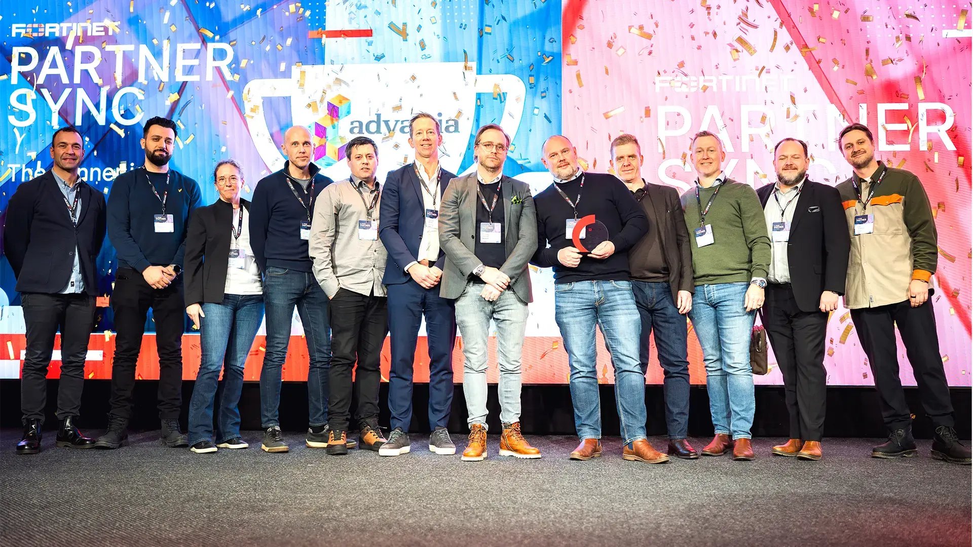 Ett team från Advania står samlade på en scen framför en färgglad bakgrund med texten ”Fortinet Partner Sync”. En person håller i ett pris, och flera bär namnskyltar runt halsen. Stämningen är stolt och festlig.