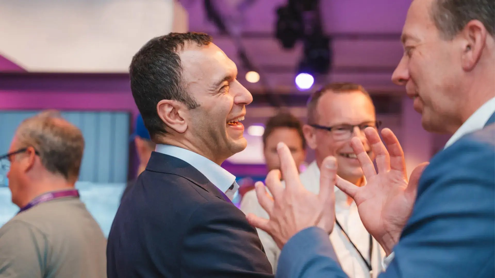 En grupp personer i kostym samtalar och skrattar i en livlig miljö med lila belysning. Fokus ligger på två män i förgrunden som uttrycker sig med händerna i ett engagerat samtal.