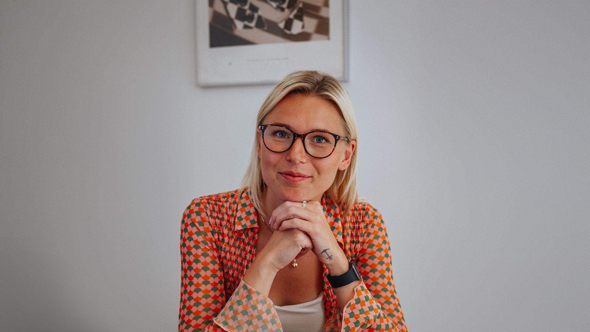 En blond kvinna med glasögon och röd-grönmönstrad skjorta sitter vid ett bord med händerna vilande under hakan. En tavla hänger på väggen bakom henne.