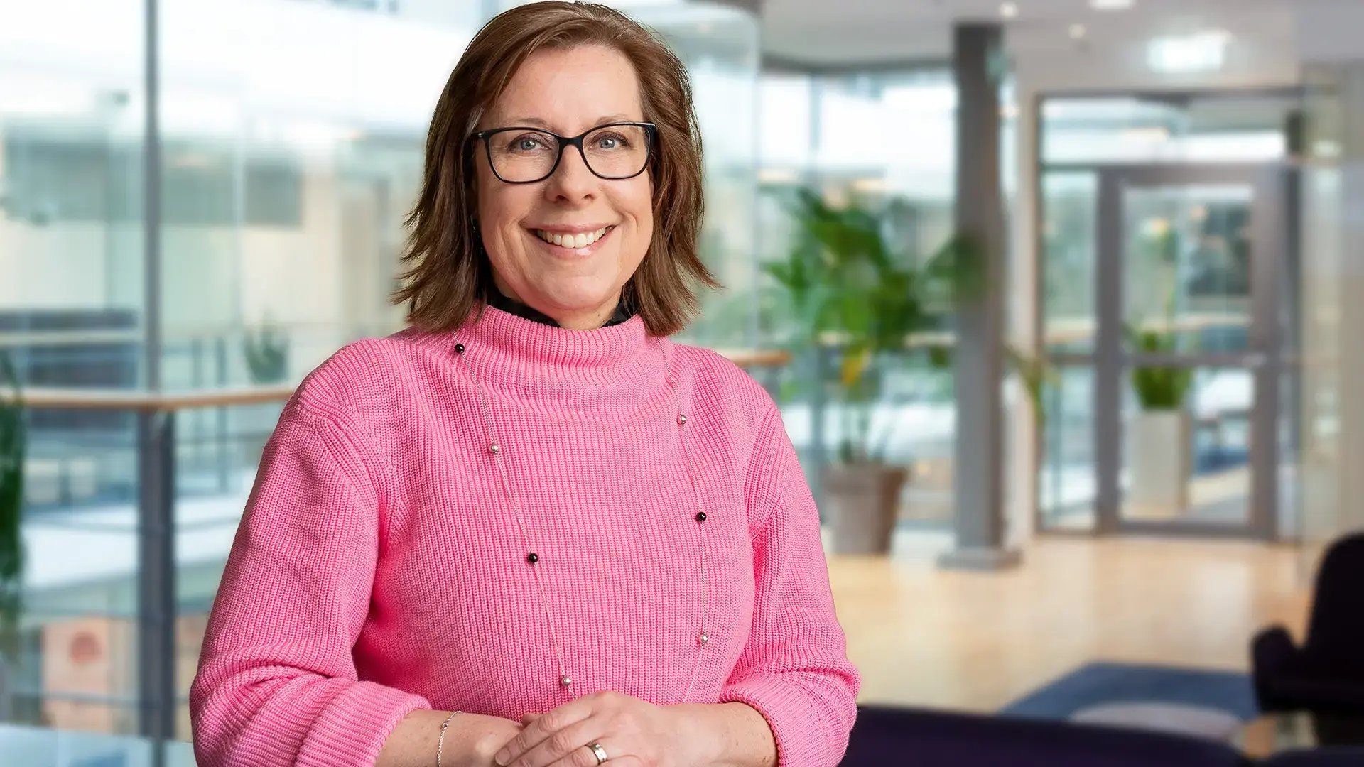 En leende kvinna med glasögon och rosa tröja står i ett ljust kontorslandskap med glasväggar och gröna växter.