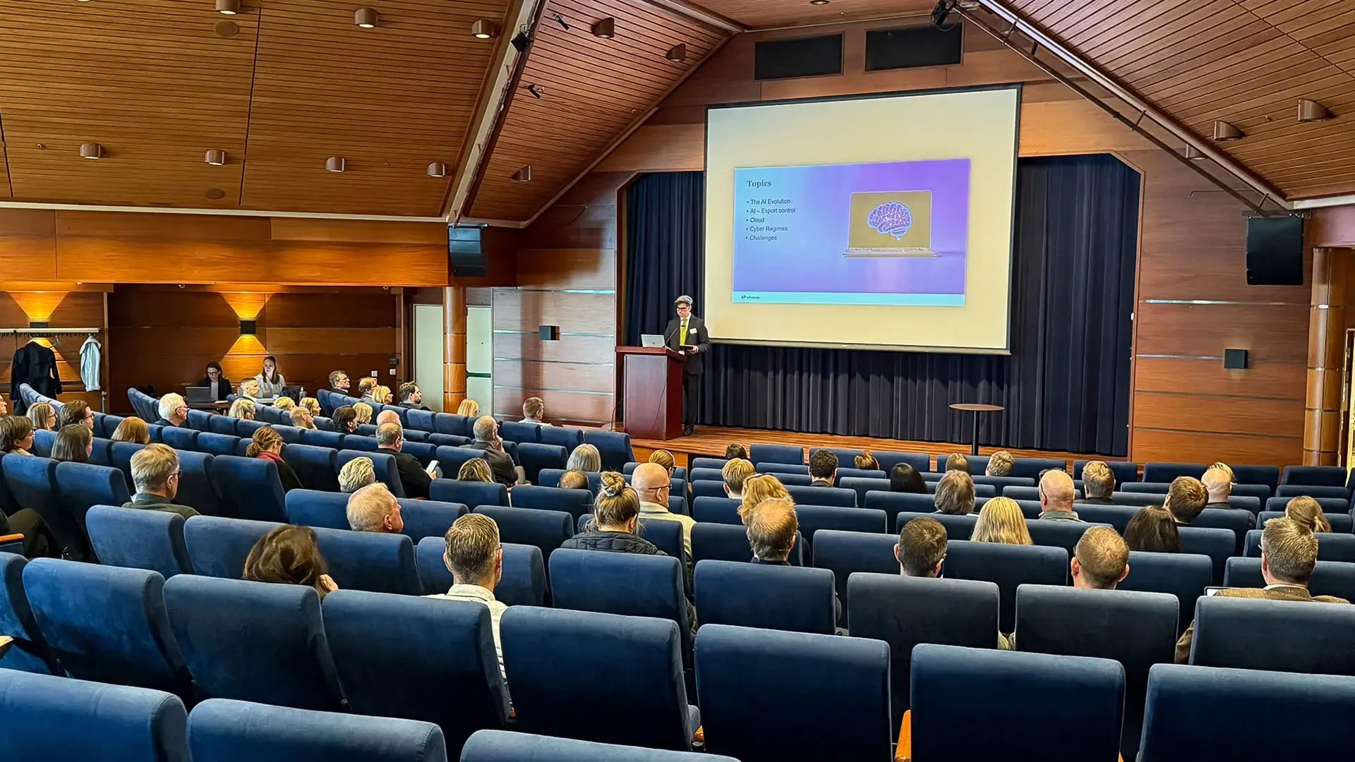 En föreläsare står vid ett podie i en träklädd sal och presenterar inför en sittande publik. På projektorduken visas en bild med rubriken ”Topics” och en illustration av en hjärna. Åhörarna sitter i blå fåtöljer och lyssnar uppmärksamt.