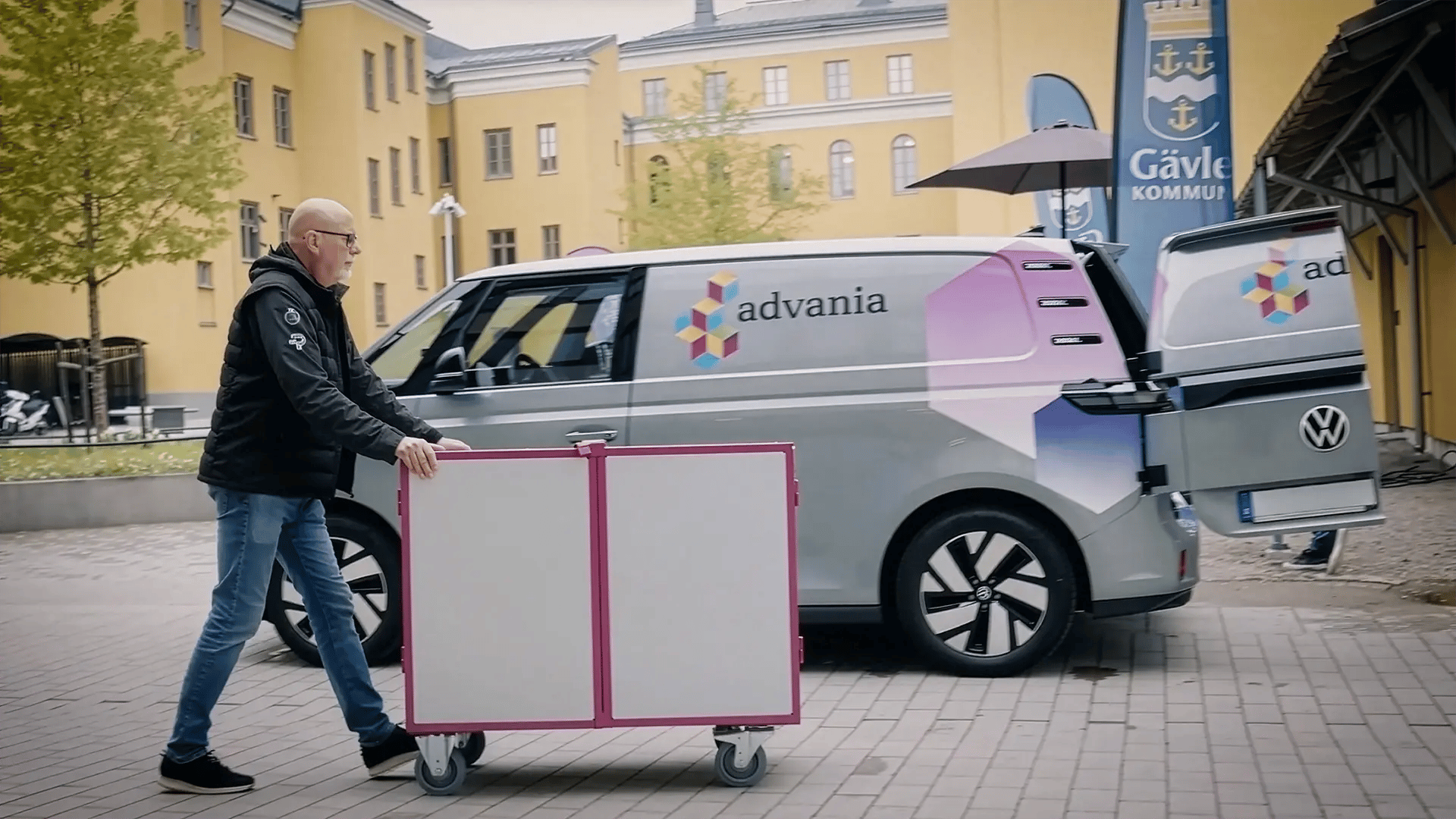 Man skjuter vagn framför Advania-bil vid gul byggnad.
